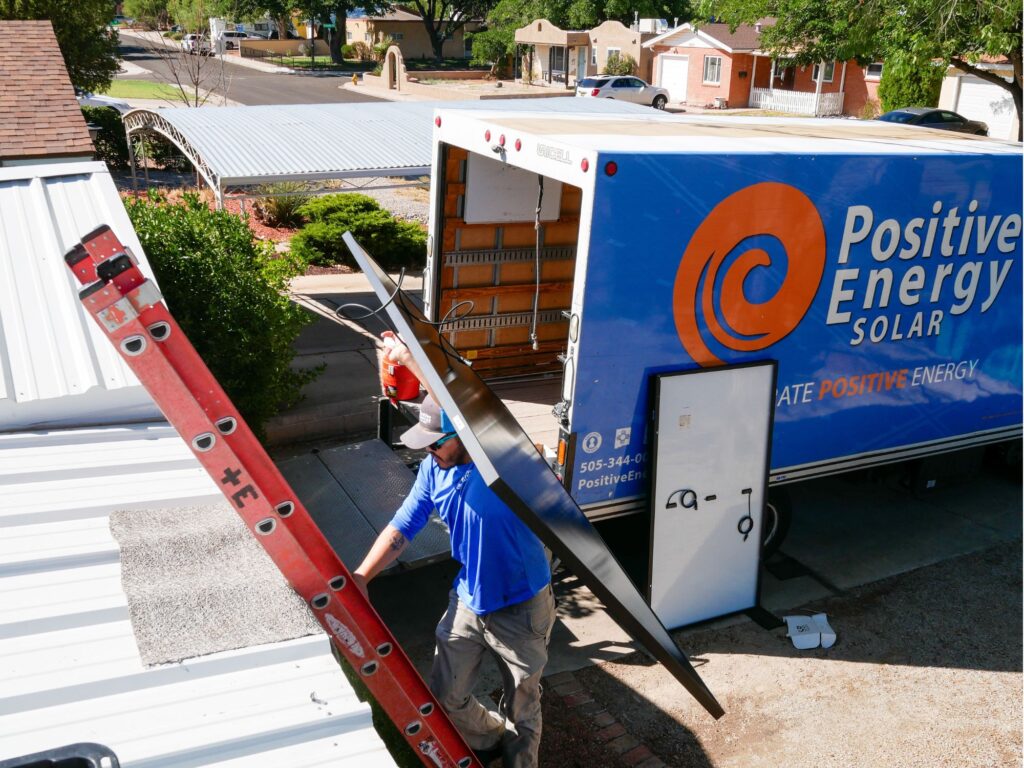 Worker carrying a solar panel up a ladder for installation. Positive Energy Solar truck in the background.